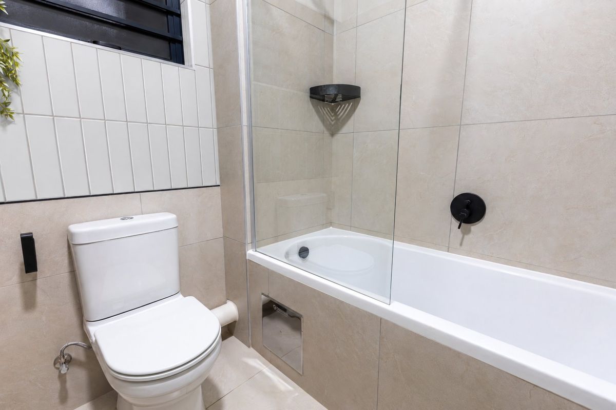 Modern bathroom featuring a white toilet, a bathtub with a glass screen, and neutral-toned wall and floor tiles.