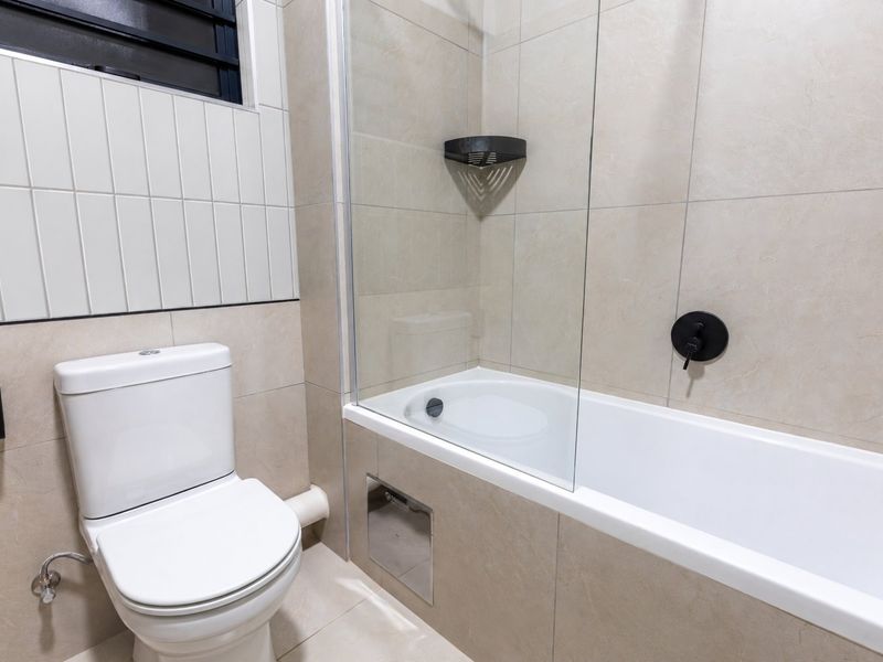 Modern bathroom featuring a white toilet, a bathtub with a glass screen, and neutral-toned wall and floor tiles.