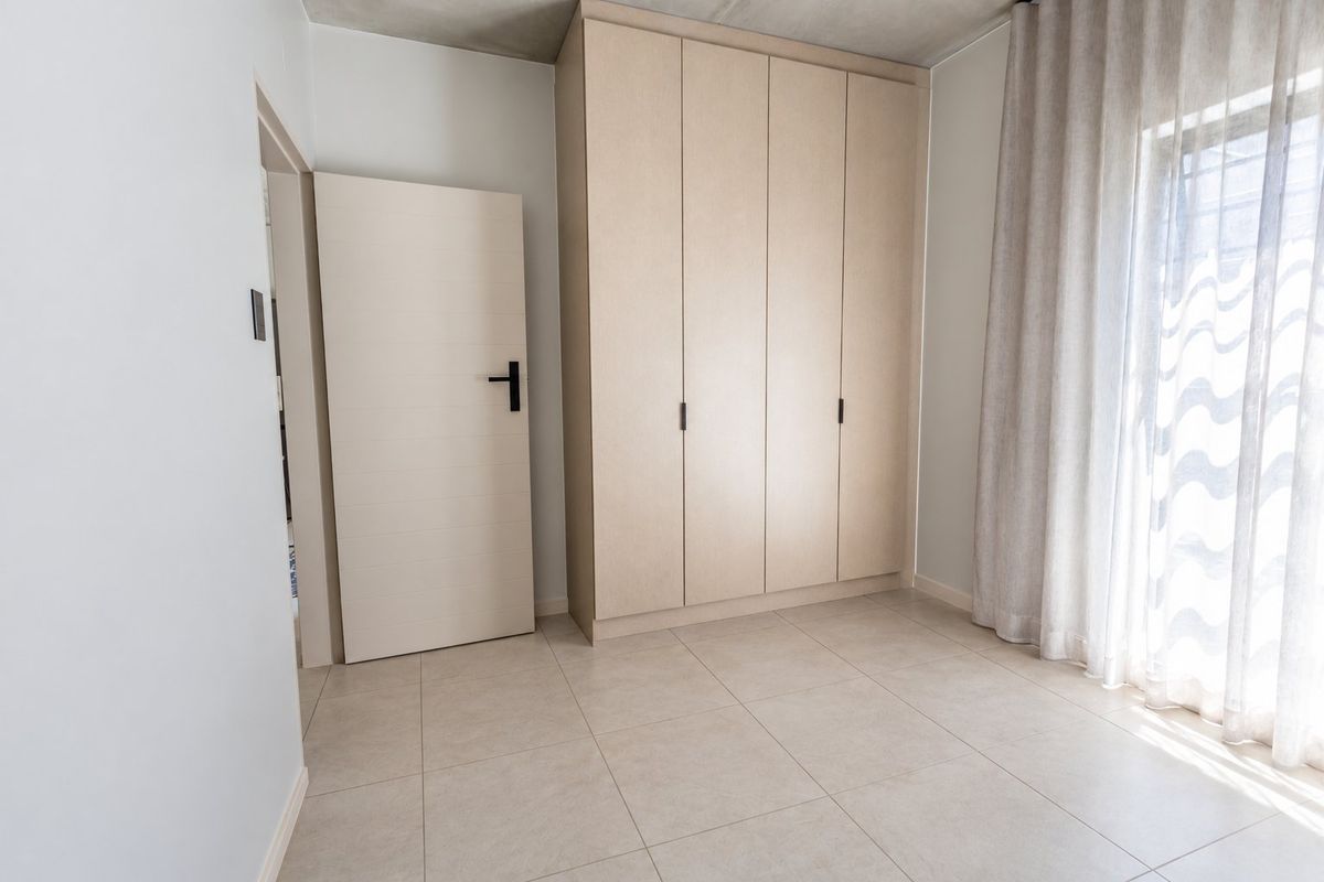 A bright, modern bedroom featuring neutral tiled flooring, a large built-in wardrobe with light wood finish, and natural light from the window.