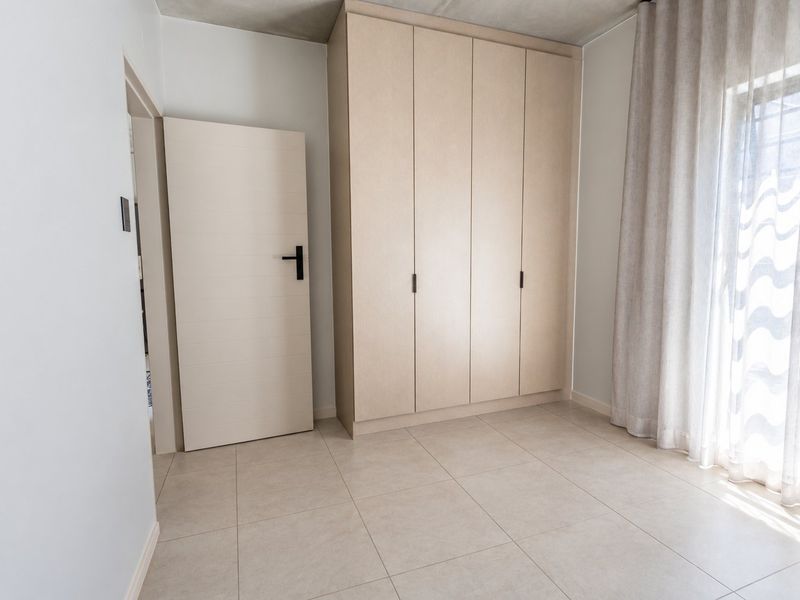 A bright, modern bedroom featuring neutral tiled flooring, a large built-in wardrobe with light wood finish, and natural light from the window.