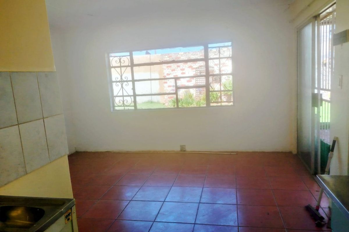 An empty open-plan living and kitchen area featuring tiled flooring, a sliding glass door, and security window bars, awaiting modernization.
