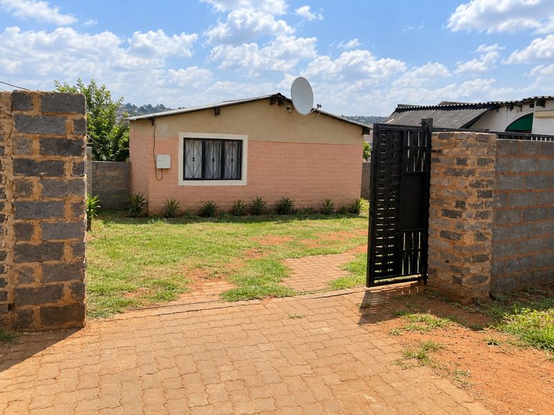 Front view of a modest house featuring a paved driveway, metal entrance gate, and a brick boundary wall with a small front garden area.