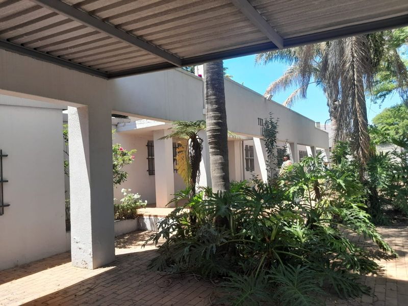 A covered outdoor patio area featuring brick paving and structural pillars, providing a shaded transition space between the building and the garden.