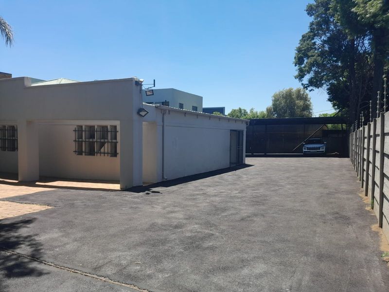 Rear exterior view of the commercial property featuring a large asphalt driveway, secure perimeter fencing, and a covered parking area.