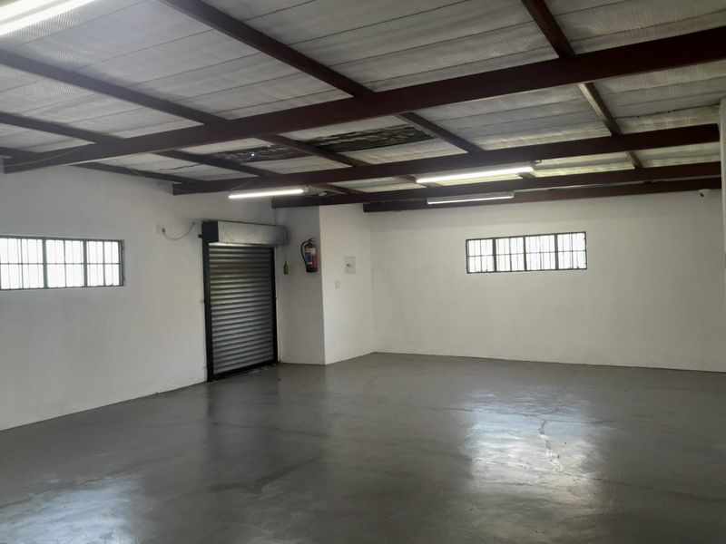 Spacious commercial utility area featuring exposed steel beams, concrete flooring, a roller shutter door, and security-barred windows, suitable for storage or workshop use.