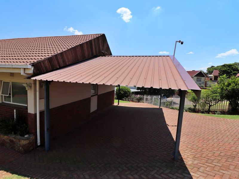 A well-maintained front exterior featuring a covered carport with a corrugated metal roof, brick paving, and a stone planter box.