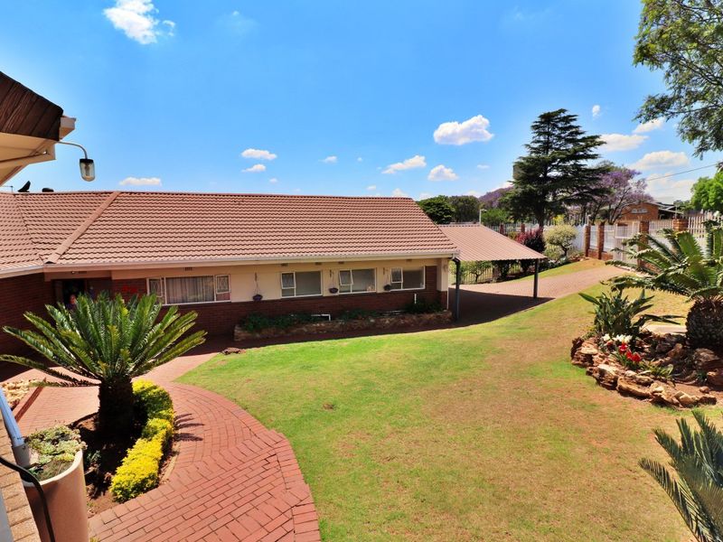 A well-maintained rear exterior view featuring a brick house with a tiled roof, a carport, paved pathways, and a landscaped garden with stone retaining walls.