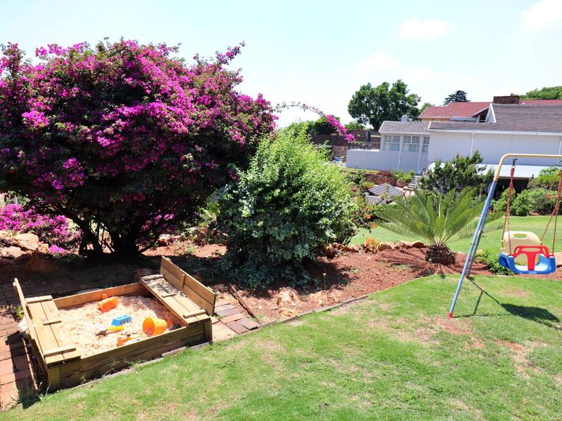A well-maintained residential garden featuring a lush lawn, mature flowering shrubs, and a dedicated play area with a sandbox.