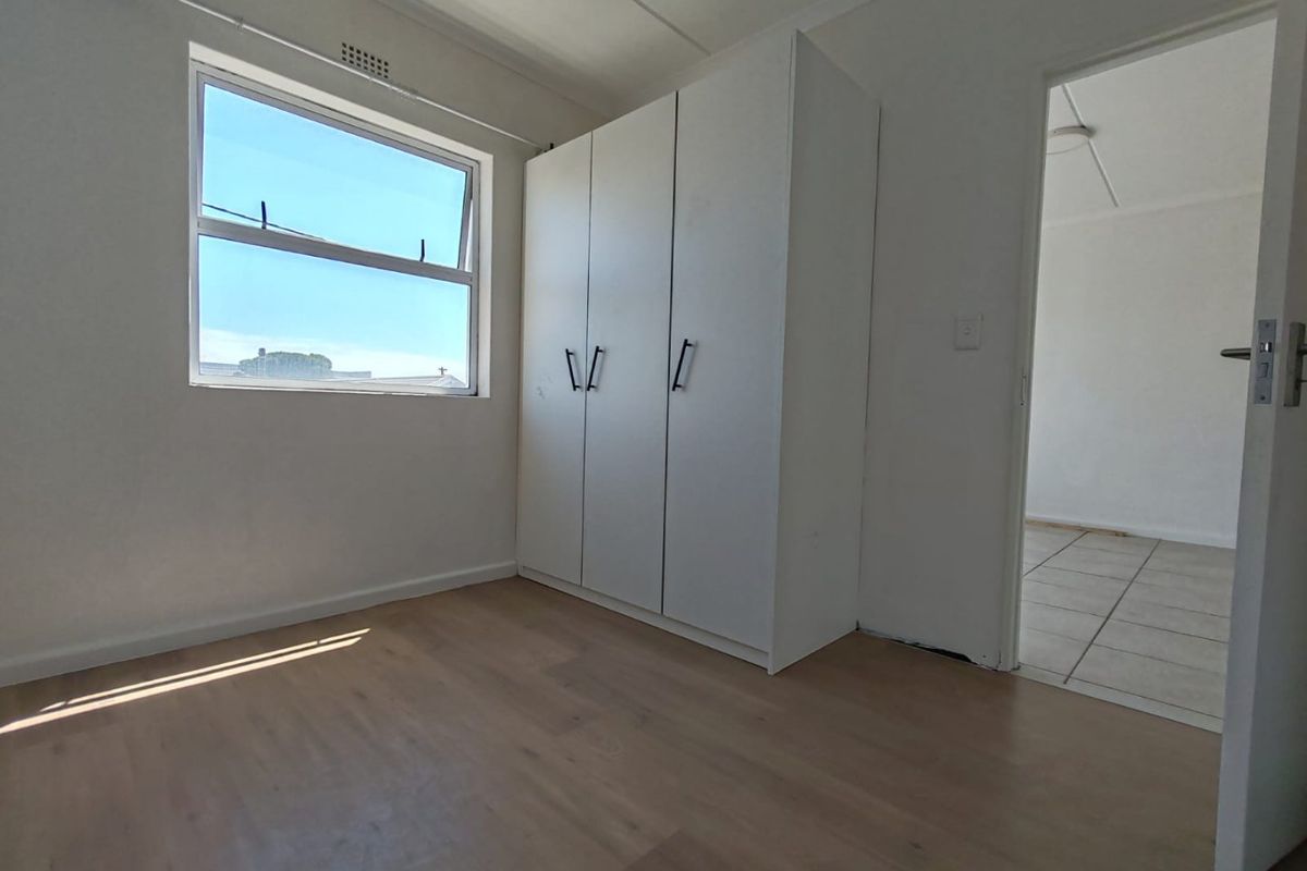 A bright, empty bedroom featuring light laminate flooring, a built-in white wardrobe, and a window providing natural light.