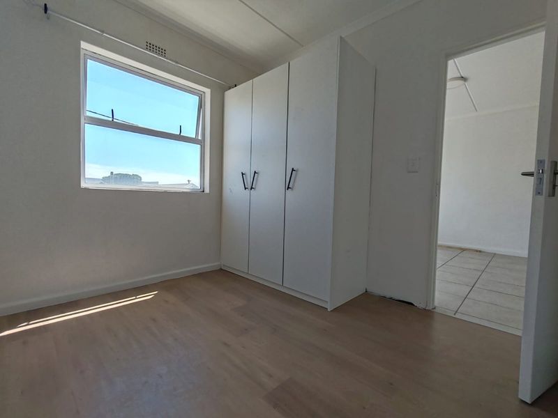 A bright, empty bedroom featuring light laminate flooring, a built-in white wardrobe, and a window providing natural light.