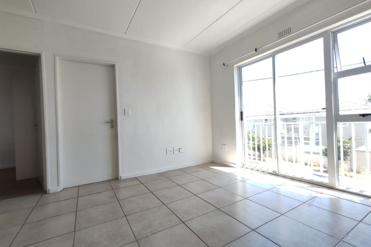 A bright, empty living area featuring neutral tiled flooring and a large sliding glass door providing natural light and access to a balcony.
