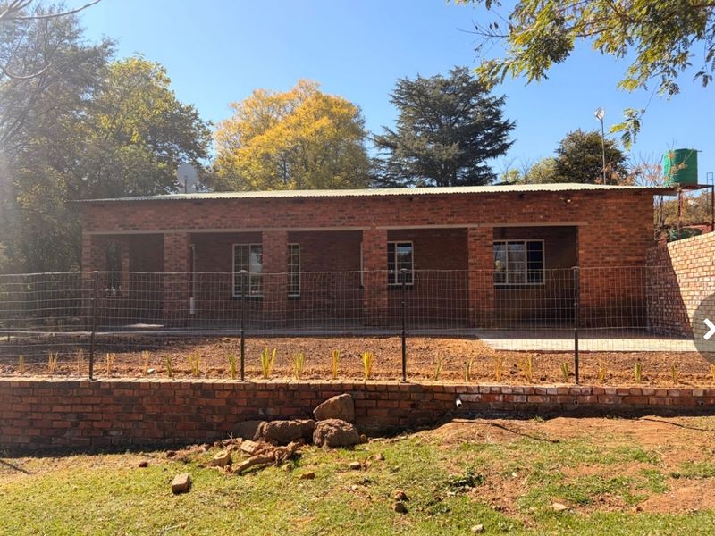Rear view of a single-story exposed brick house featuring a covered patio, corrugated roof, and a tiered garden area with retaining wall.