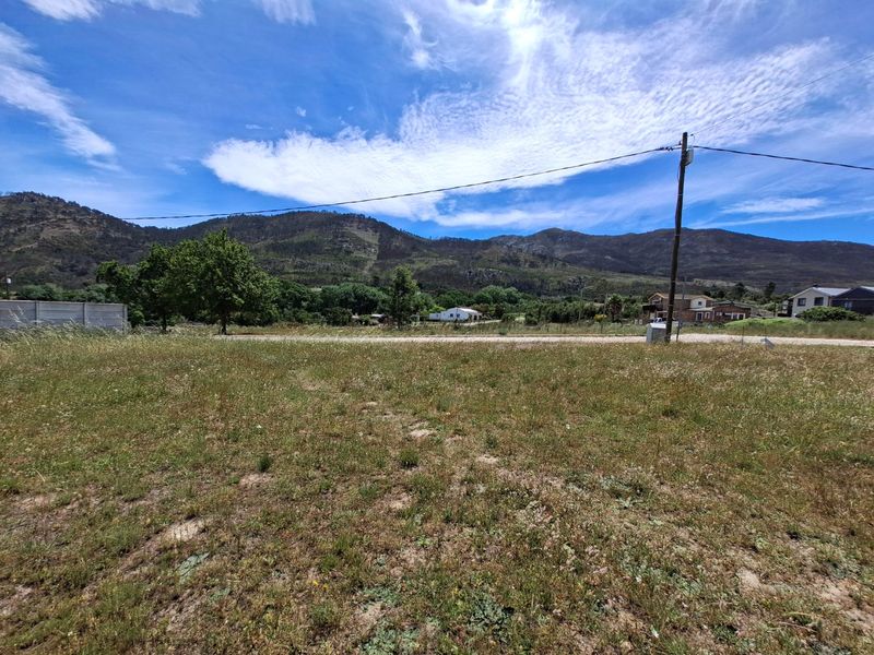 A vacant residential plot featuring natural grass terrain, a boundary wall, and scenic mountain views under a clear blue sky.