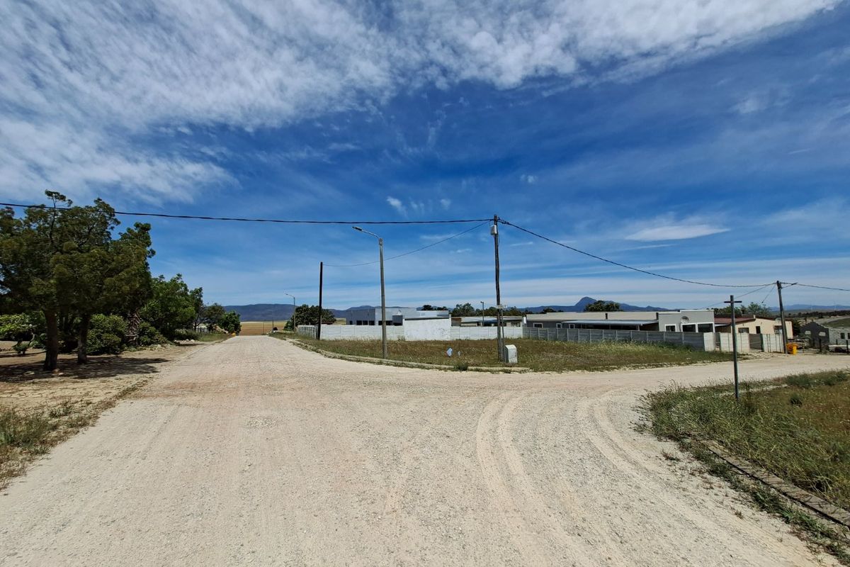A street-level view of a vacant residential plot featuring a gravel road, utility infrastructure, and surrounding boundary walls.