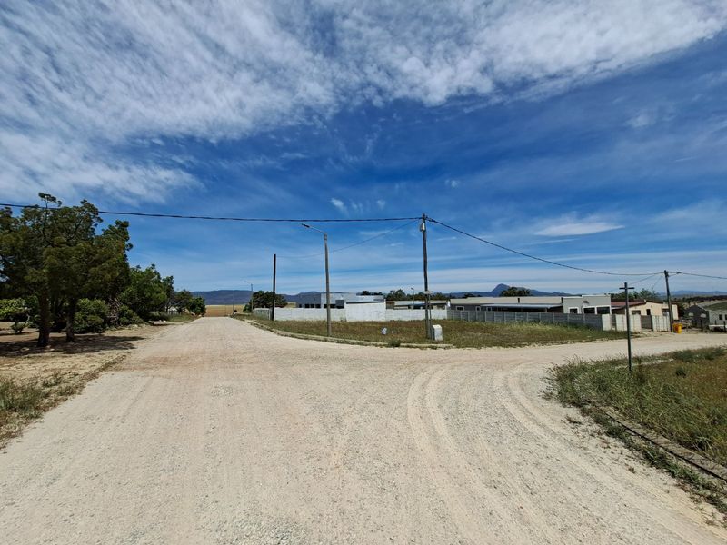 A street-level view of a vacant residential plot featuring a gravel road, utility infrastructure, and surrounding boundary walls.