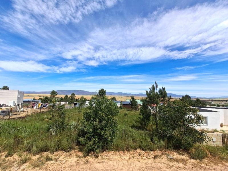 A wide-angle view of the residential land plot featuring natural vegetation, surrounding structures, and distant mountain scenery under a clear blue sky.