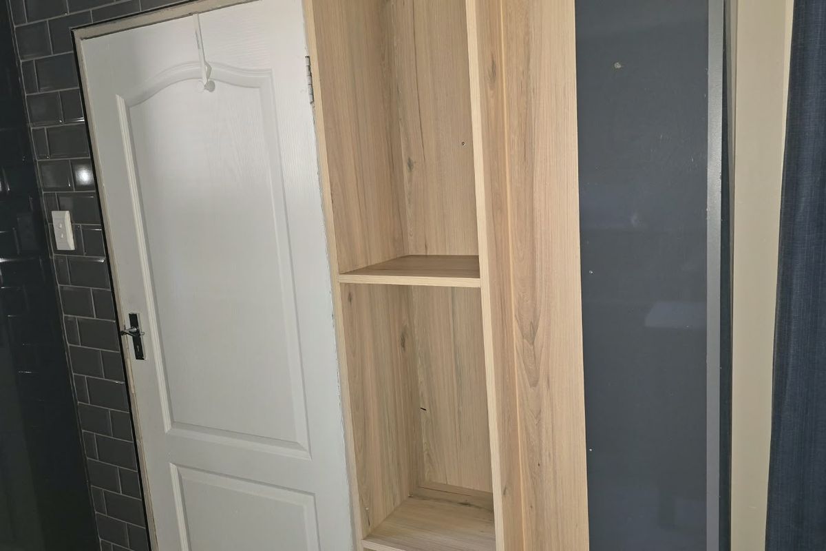 A bathroom interior featuring built-in light wood shelving, dark subway tile wall cladding, and a white panel door with visible unfinished trim work.