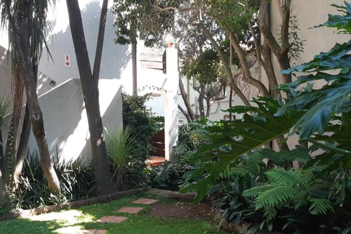 A view of the rear exterior area featuring a landscaped garden, mature trees, and a paved pathway leading towards the building entrance.