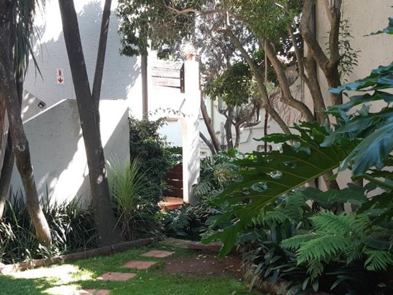 A view of the rear exterior area featuring a landscaped garden, mature trees, and a paved pathway leading towards the building entrance.