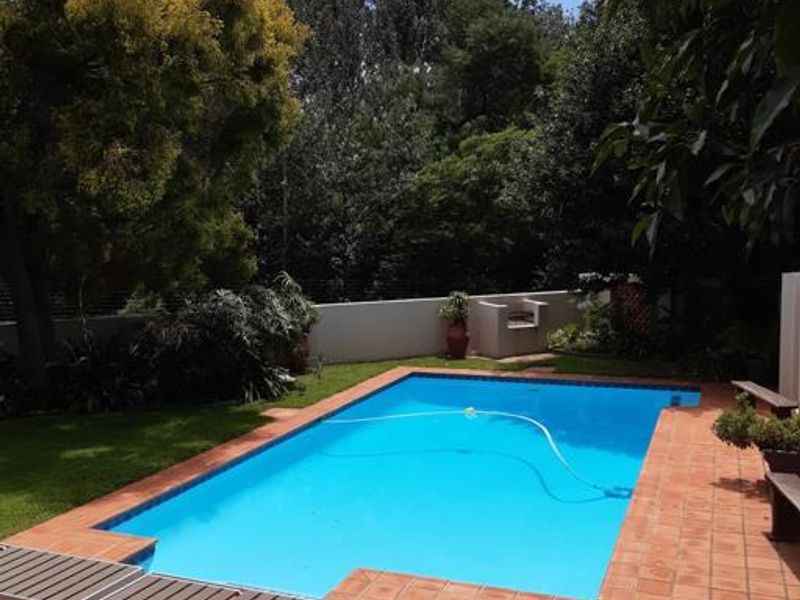 A well-maintained outdoor swimming pool area featuring a tiled surround, built-in braai, and established garden surroundings.