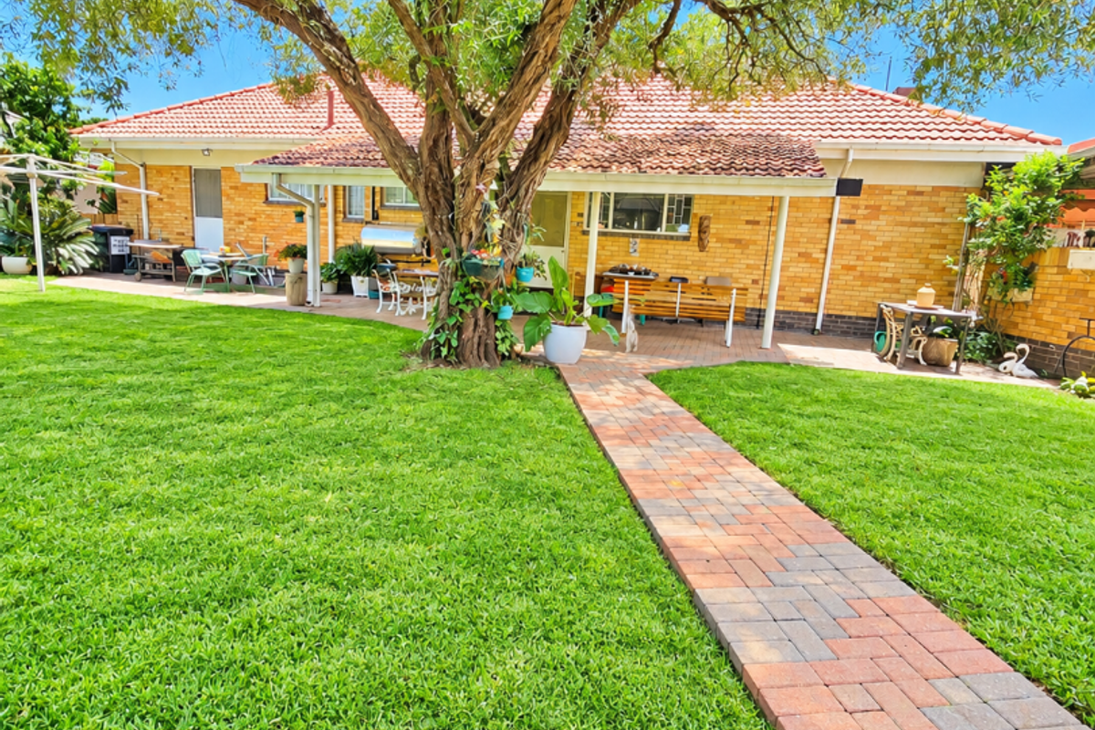 A well-maintained rear garden featuring a paved walkway leading to a covered patio area with a brick facade and tiled roof.