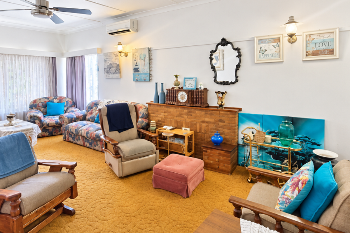 A spacious living room featuring a brick fireplace, ceiling fan, and wall-mounted air conditioning unit, finished with wall-to-wall carpeting.