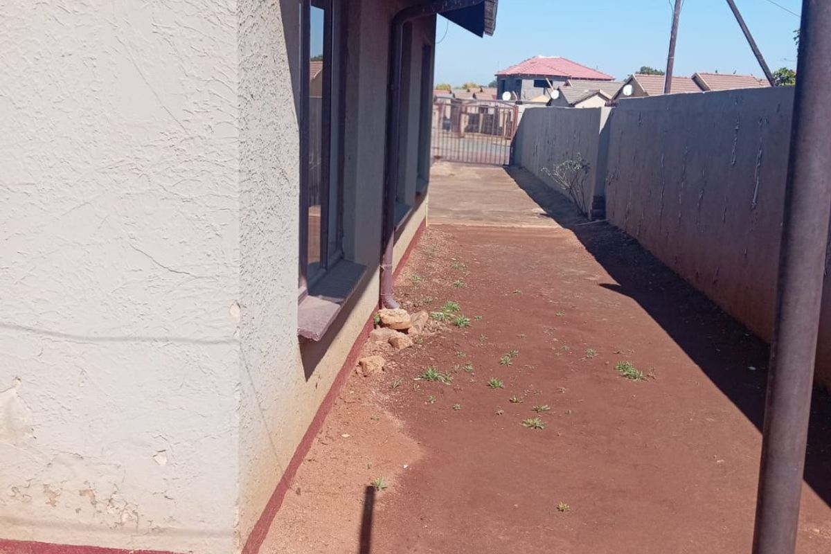 Side exterior view of the property showing a narrow pathway, perimeter wall, and basic building finishes requiring maintenance.