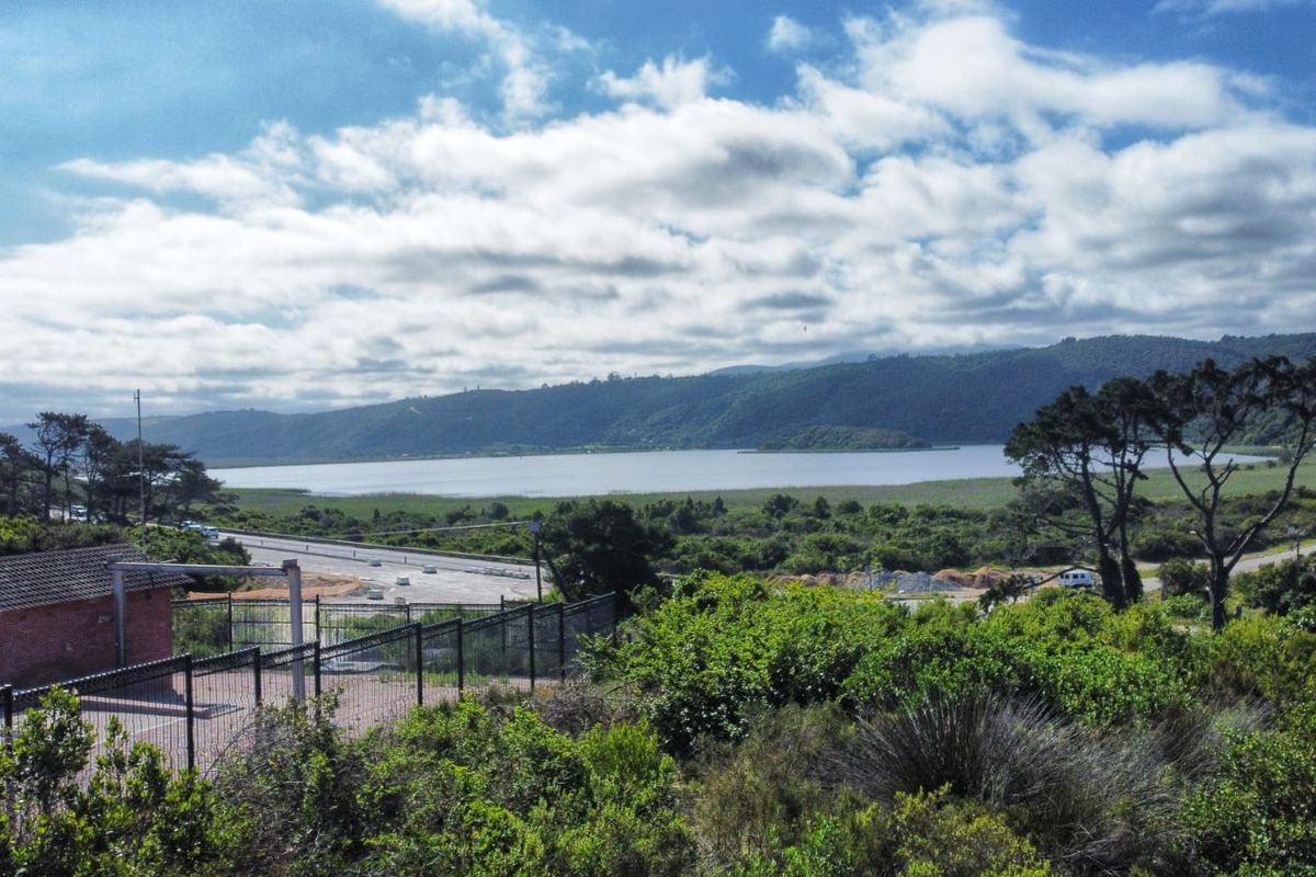 A scenic elevated view overlooking a lake, surrounding hills, and local road infrastructure, featuring a small brick utility building and perimeter fencing in the foreground.