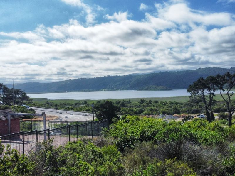 A scenic elevated view overlooking a lake, surrounding hills, and local road infrastructure, featuring a small brick utility building and perimeter fencing in the foreground.