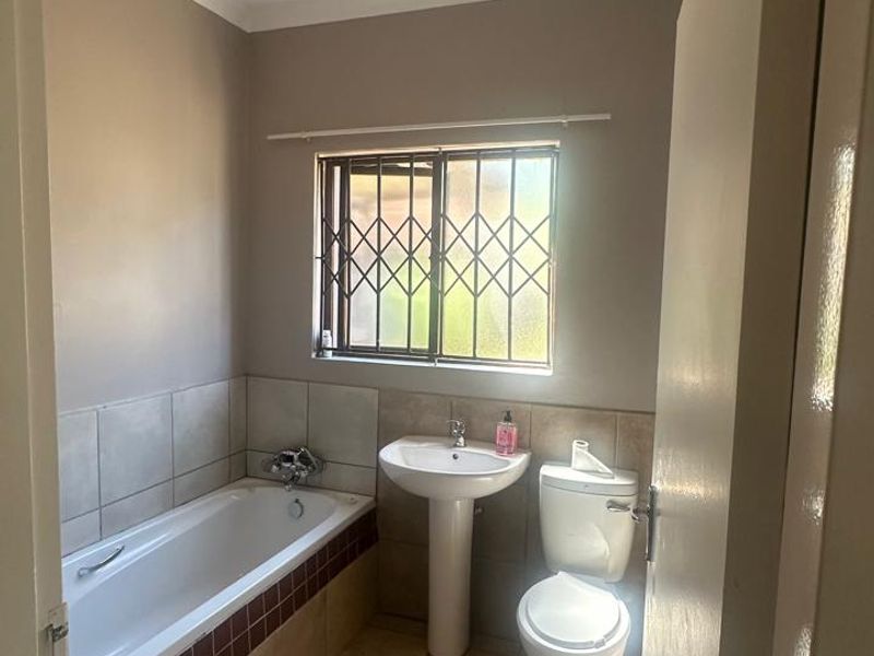 A functional bathroom featuring a bathtub, pedestal basin, and toilet with neutral tiled finishes and a security-fitted window.