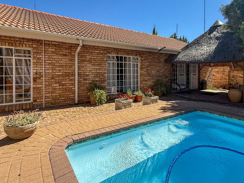 A well-maintained outdoor pool area featuring a swimming pool, brick paving, and a traditional thatched lapa, set against a face brick townhouse exterior.
