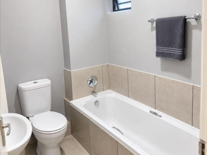 A clean and well-maintained bathroom featuring a bathtub, toilet, and washbasin with neutral tile finishes and a small louvered window.