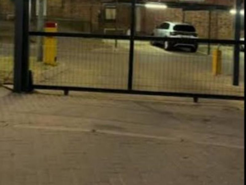 View of the apartment complex entrance featuring a security gate, paved driveway, and covered parking area.