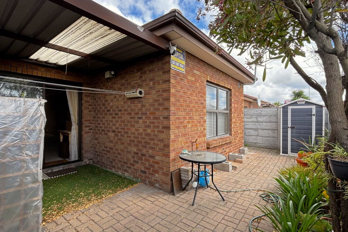 Rear exterior view featuring a brick facade, covered patio area, paved courtyard, and a storage shed within a fenced garden space.