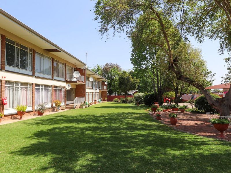 A well-maintained rear exterior view of an apartment complex featuring a large green lawn, mature trees, and a paved patio area.