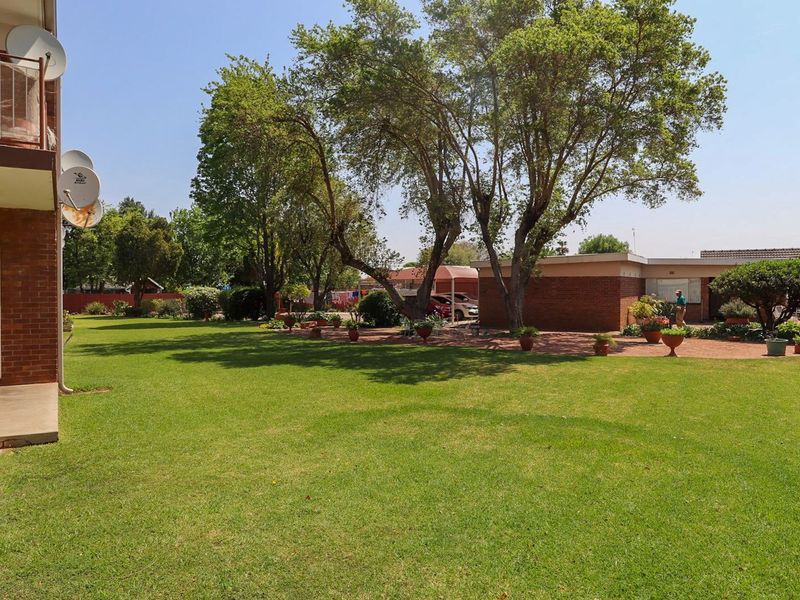 A view of the rear exterior of the apartment complex, featuring a well-maintained lawn, paved patio area, and mature trees.