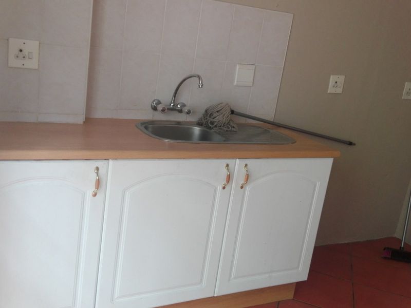 A functional kitchen area featuring white cabinetry, a stainless steel sink with mixer tap, laminate countertops, and terracotta floor tiles.