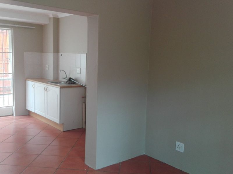 An open-plan living area featuring tiled flooring and a compact kitchen with built-in cabinetry, sink, and access to an exterior security gate.