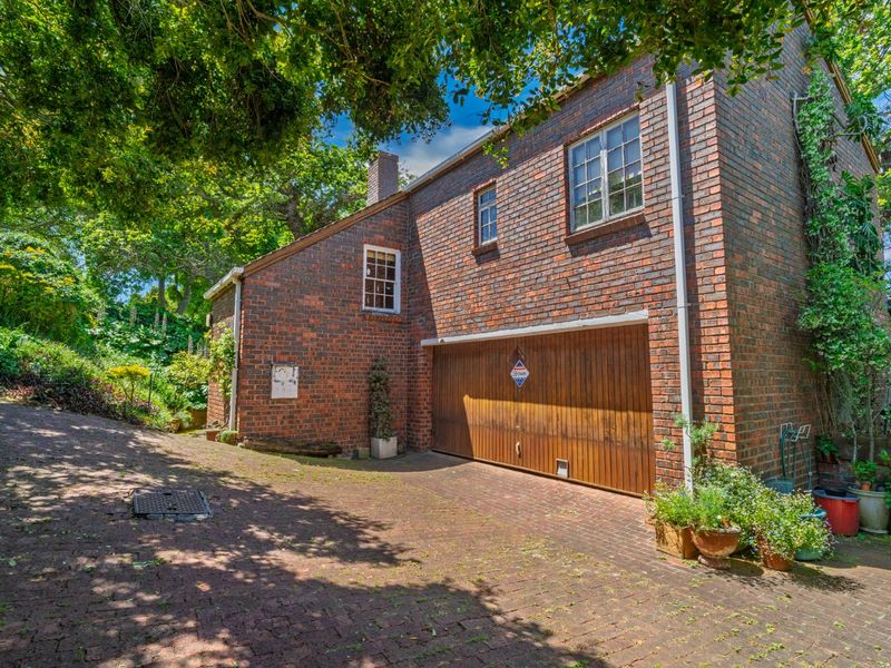 A charming double-story brick home featuring a wooden garage door, paved driveway, and established garden surroundings.