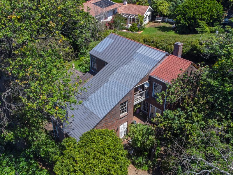Aerial view of a brick house featuring a multi-pitched roof, chimney, and balcony, surrounded by mature trees in a lush garden setting.