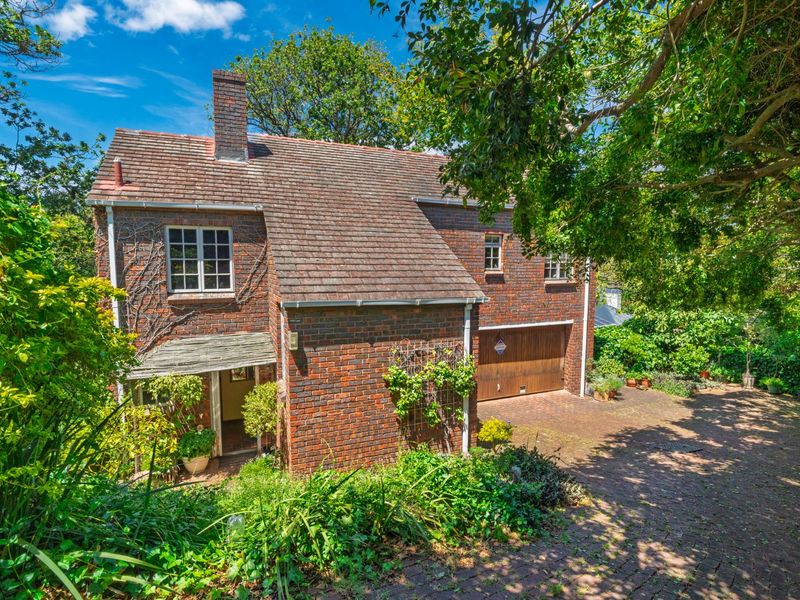 A charming two-story brick house featuring a tiled roof, integrated garage, and a paved driveway, surrounded by mature garden landscaping.