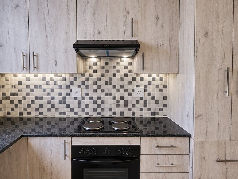 Modern kitchen featuring light wood-grain cabinetry, dark granite countertops, a mosaic tile backsplash, and integrated electric cooking appliances with under-cabinet lighting.