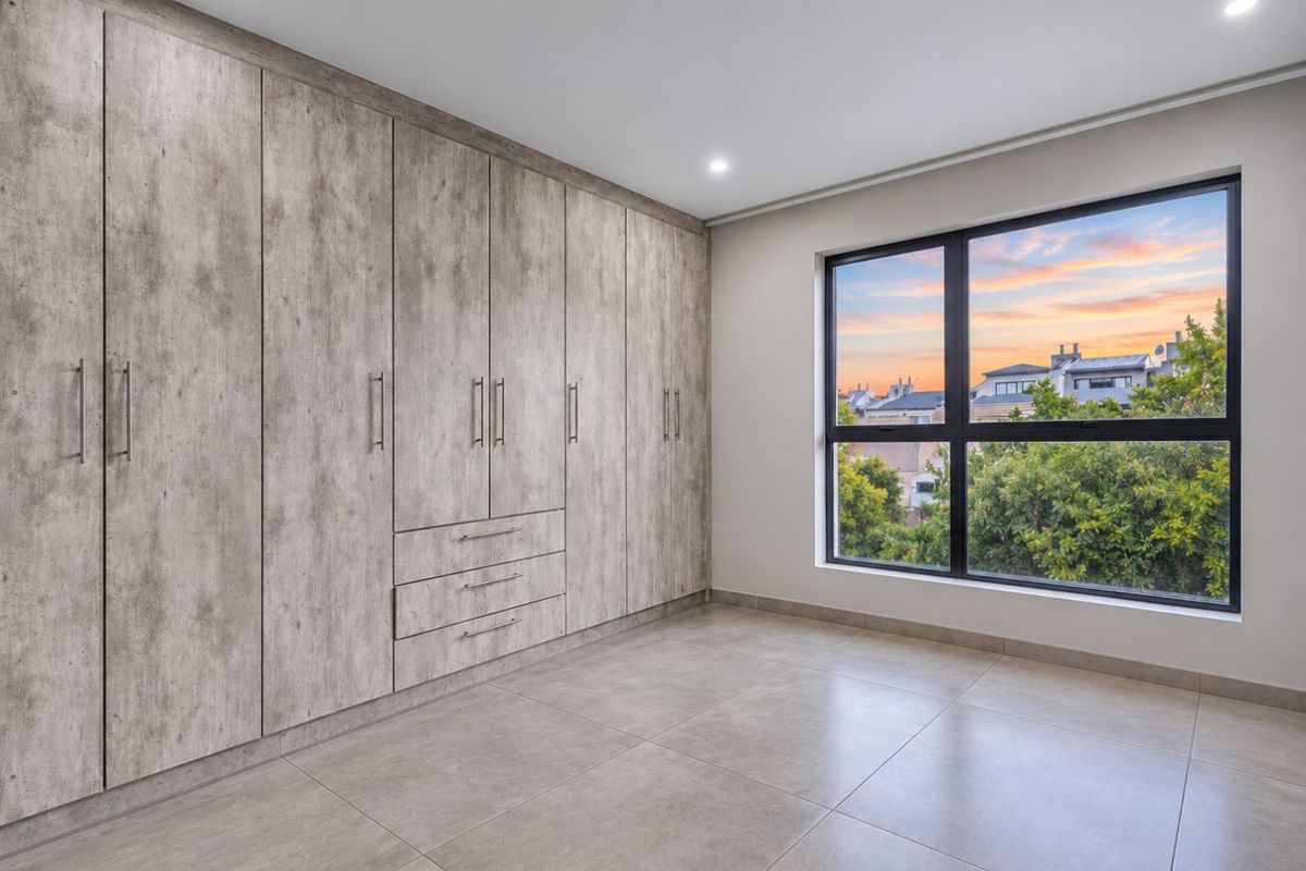 A bright, modern bedroom featuring extensive built-in cupboards with a wood-grain finish, large aluminium-framed windows, and neutral tiled flooring.