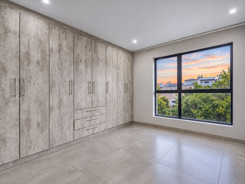 A bright, modern bedroom featuring extensive built-in cupboards with a wood-grain finish, large aluminium-framed windows, and neutral tiled flooring.