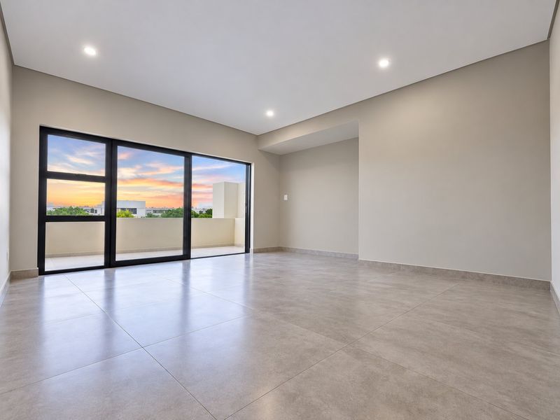 A bright, modern, and spacious living area featuring large sliding glass doors that open onto a balcony, finished with neutral tiles and recessed lighting.