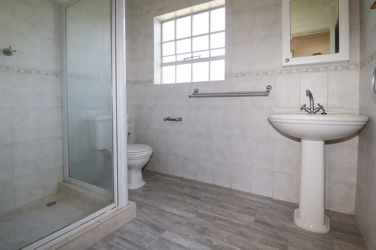 A clean, functional bathroom featuring a pedestal washbasin, a glass-enclosed shower, and a toilet, finished with neutral wall and floor tiling.