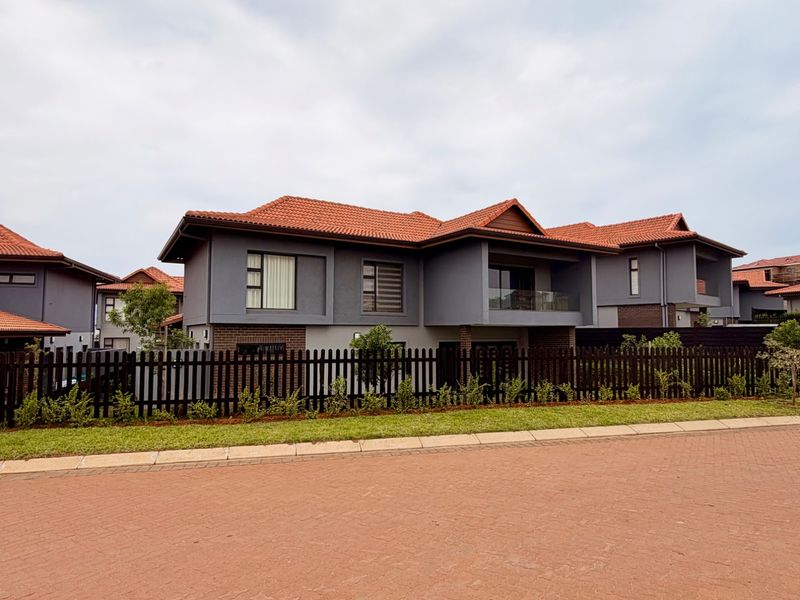 A modern double-story residential property featuring a tiled roof, dark grey facade, and a private balcony with a glass balustrade, set behind a picket fence.
