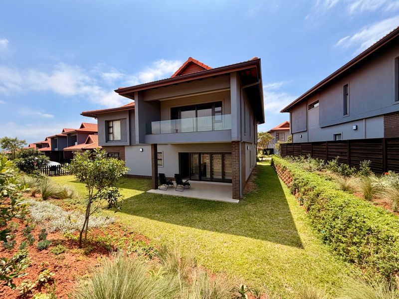 Rear view of a modern two-story house featuring a paved patio, glass-balustrade balcony, and a well-maintained landscaped garden.