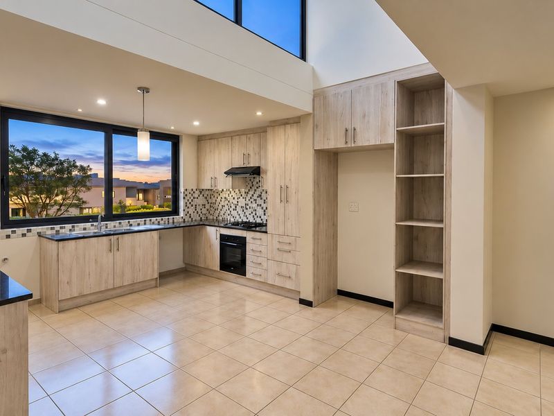 Modern kitchen featuring light wood-grain cabinetry, stone countertops, integrated appliances, and a double-height ceiling with ample natural light.