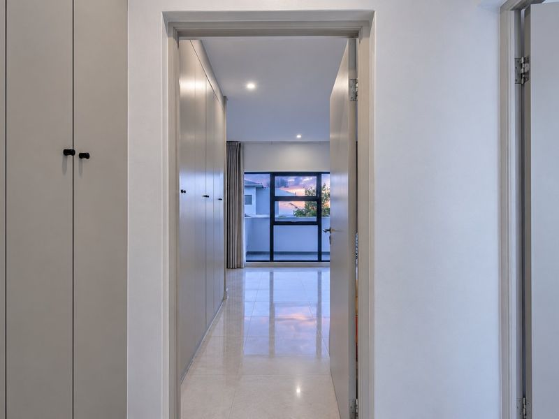 A bright, modern hallway featuring extensive built-in cabinetry, polished tiled flooring, and recessed lighting, leading towards a window with an exterior view.
